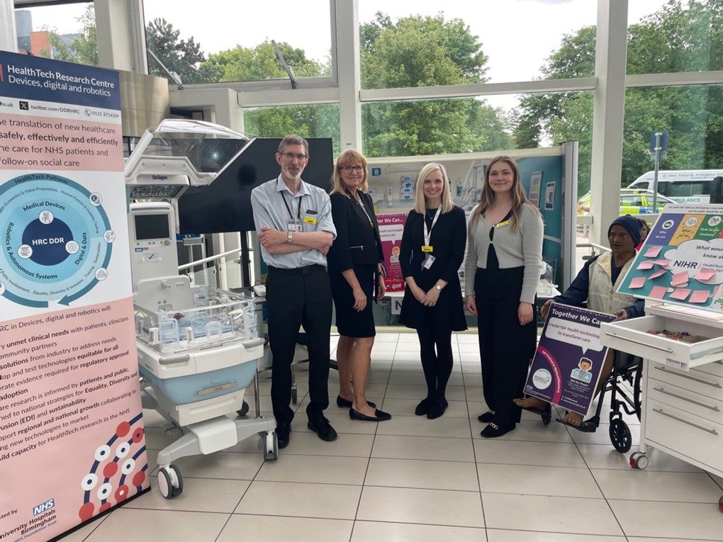Members of the HRC DDR team attending the UHB Research Showcase standing by their exhibition stand which contains banner stands and some medical equipment and a dummy patient in a wheelchair